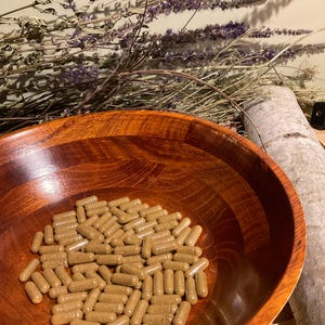 May include: A collection of light brown, cylindrical capsules resting inside a polished, dark-toned wooden bowl. The bowl features a visible wood grain pattern and a smooth, rounded interior surface, containing numerous identical supplement capsules.