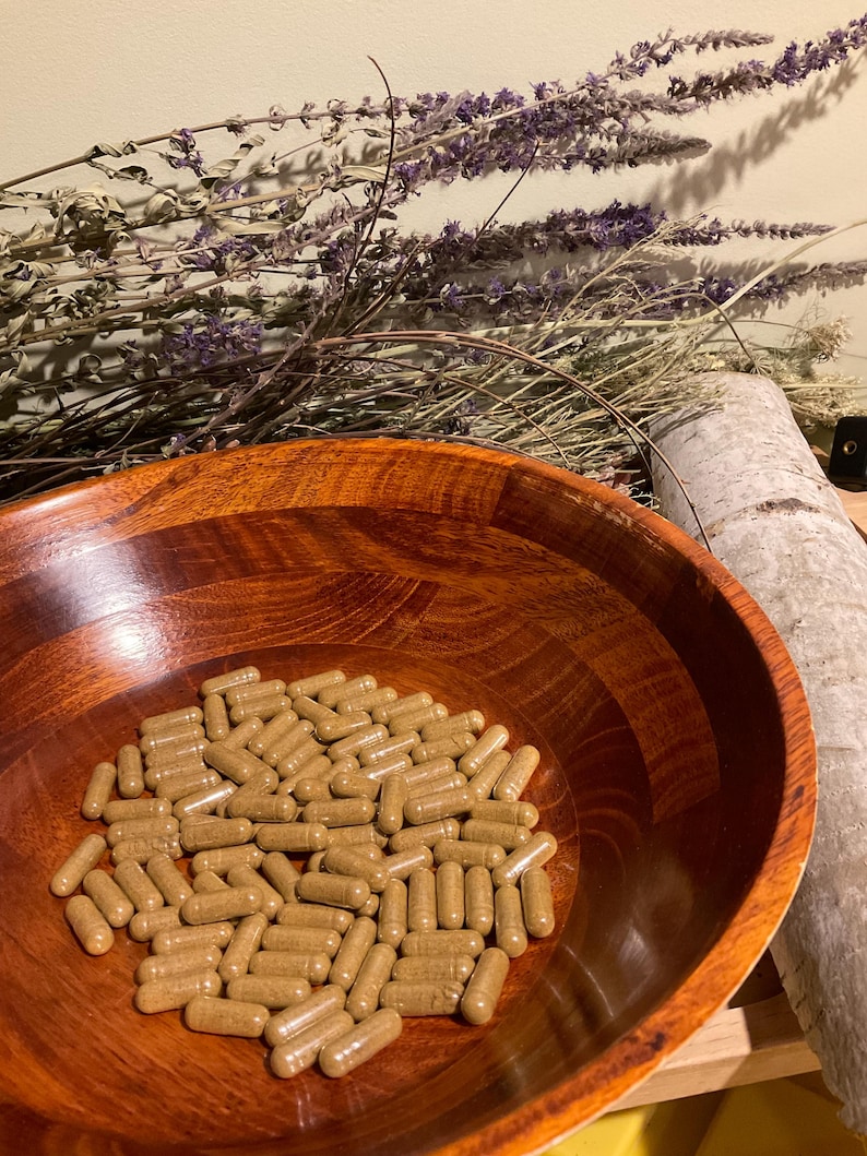 May include: A collection of light brown, cylindrical capsules resting inside a polished, dark-toned wooden bowl. The bowl features a visible wood grain pattern and a smooth, rounded interior surface, containing numerous identical supplement capsules.