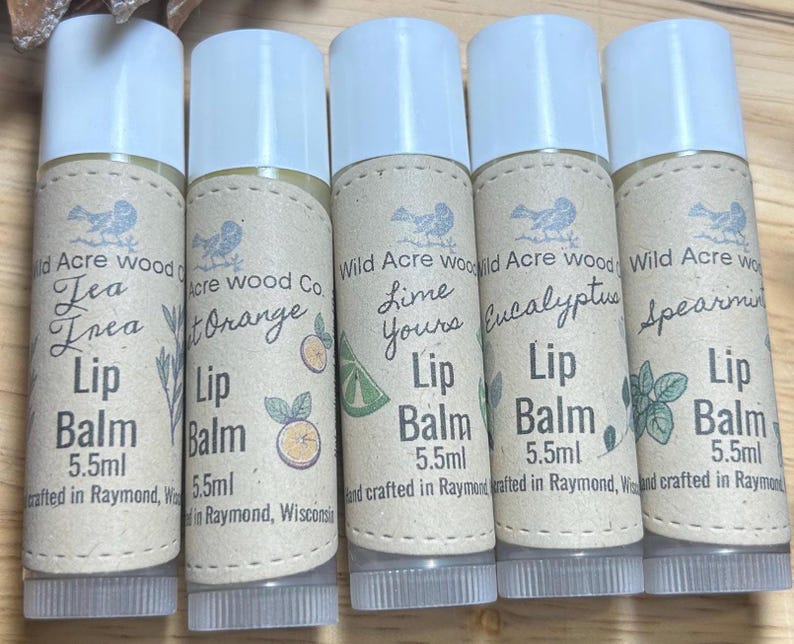 all five new lip balms lined in a row on a wooden surface in the background is a few eastern white pine, pinecones. New lip balm essential oil scents! Sweet orange, eucalyptus, Teatree, lime, spearmint. all 100% natural and organic.