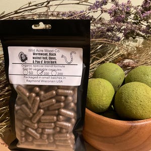 May include: A black pouch filled with brown capsules, labeled "Wild Acre Wood Co." with text including "Wormwood, Black walnut hull, Clove, & Pau d' Arco bark." Beside it, a wooden bowl holds several green, round fruits. Dried purple flowers are in the background.