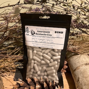 May include: A black pouch of Bentonite Clay capsules from Wild Acre Wood Co. The label reads "FOOD GRADE" and "Made in the USA." The pouch contains size 00 vegetable capsules. The background includes dried plants and a pine cone.