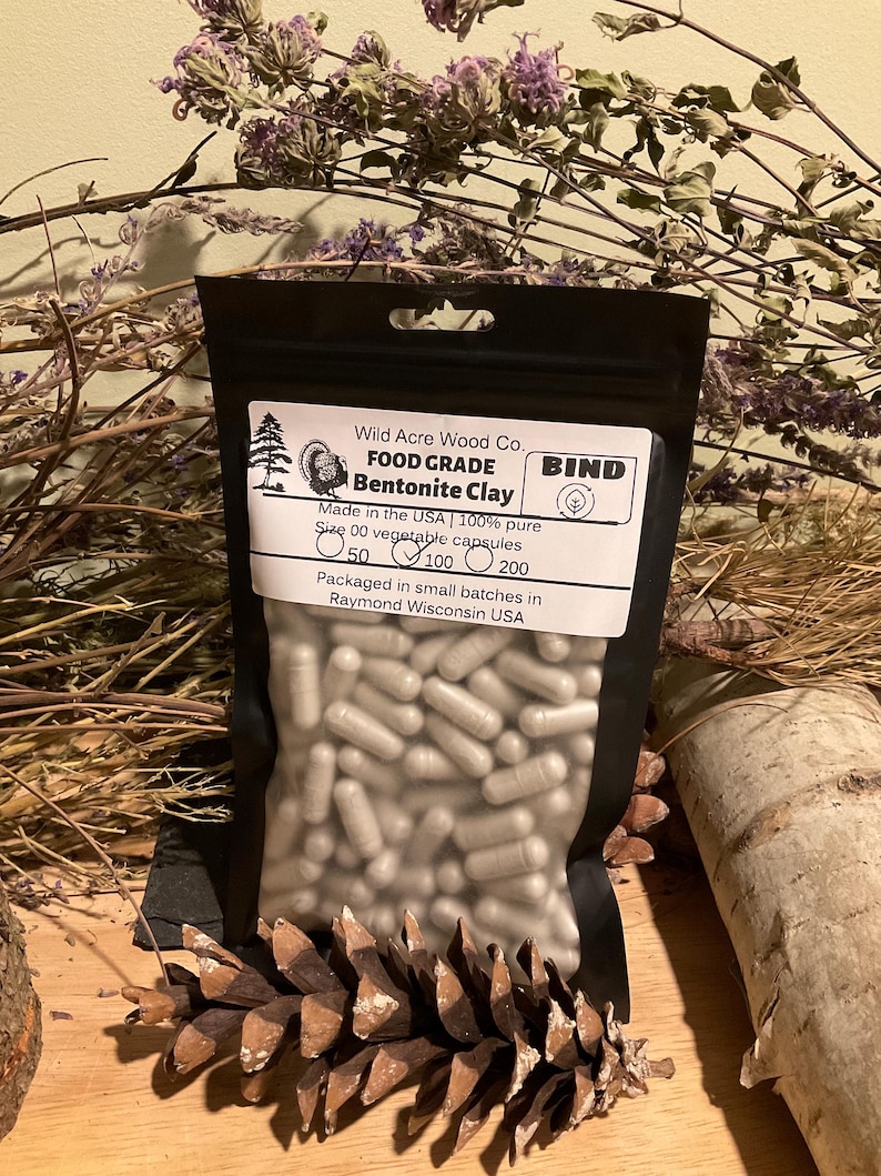 May include: A black pouch of Bentonite Clay capsules from Wild Acre Wood Co. The label reads "FOOD GRADE" and "Made in the USA." The pouch contains size 00 vegetable capsules. The background includes dried plants and a pine cone.