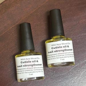 May include: Two 7 ml bottles of cuticle oil and nail strengthener by Wild Acre Wood Co. The bottles are clear glass with black caps and yellow liquid inside. The labels are white with black text and list the ingredients: grapeseed oil, sweet almond oil, vitamin E, lavender essential oil, geranium essential oil, and incense essential oil. The labels also state that the product is not evaluated by the FDA.