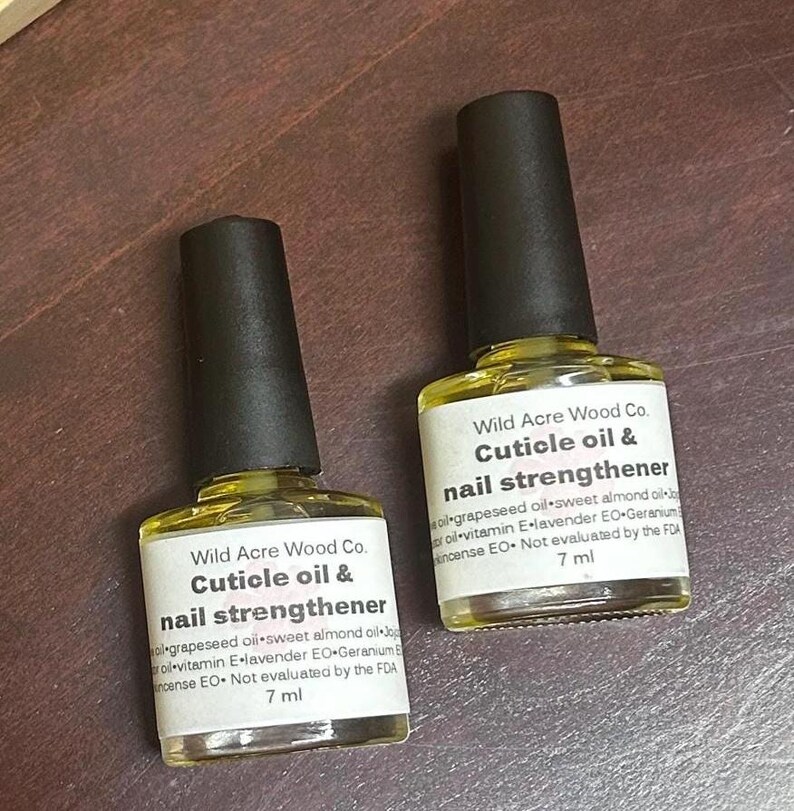 May include: Two 7 ml bottles of cuticle oil and nail strengthener by Wild Acre Wood Co. The bottles are clear glass with black caps and yellow liquid inside. The labels are white with black text and list the ingredients: grapeseed oil, sweet almond oil, vitamin E, lavender essential oil, geranium essential oil, and incense essential oil. The labels also state that the product is not evaluated by the FDA.