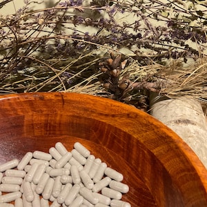 May include: A wooden bowl filled with numerous gray capsules. The bowl has a rich, reddish-brown color and a smooth, polished finish. Dried herbs and a pine cone are in the background, creating a natural, earthy aesthetic.