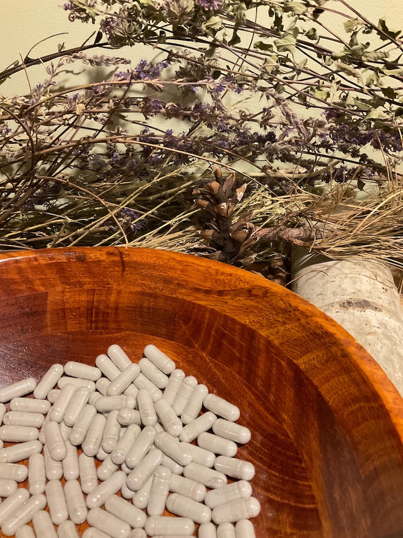 May include: A wooden bowl filled with numerous gray capsules. The bowl has a rich, reddish-brown color and a smooth, polished finish. Dried herbs and a pine cone are in the background, creating a natural, earthy aesthetic.