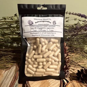 May include: A black resealable pouch containing a bulk quantity of beige capsules labeled "Ashwagandha" with the text "Wild Acre Wood Co. Withania somnifera USDA organic | GMP certified Size 00 vegetable capsules 50 100 200 Packaged in small batches in Raymond Wisconsin USA".
