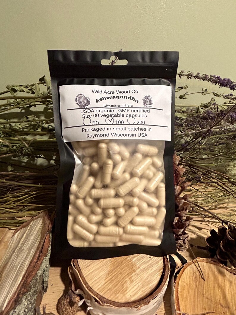 May include: A black resealable pouch containing a bulk quantity of beige capsules labeled "Ashwagandha" with the text "Wild Acre Wood Co. Withania somnifera USDA organic | GMP certified Size 00 vegetable capsules 50 100 200 Packaged in small batches in Raymond Wisconsin USA".