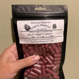 May include: A black bag filled with maroon capsules, labeled "Organic Beetroot" and "USDA Organic gluten-free." The bag also indicates "Size 00 vegetable capsules" and the quantity of 100 capsules. The text "Wild Acre Wood Co." is also visible.