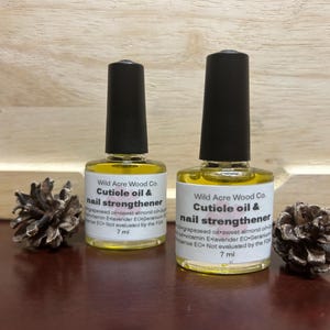 May include: Two 7 ml bottles of cuticle oil and nail strengthener by Wild Acre Wood Co. The bottles are clear glass with a yellow liquid inside. The labels are white with black text. The bottles are sitting on a wooden surface.