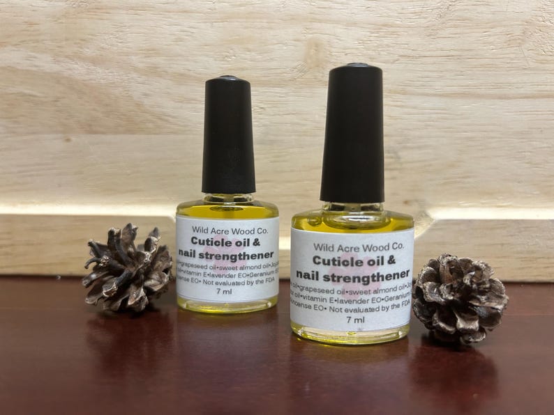 May include: Two 7 ml bottles of cuticle oil and nail strengthener by Wild Acre Wood Co. The bottles are clear glass with a yellow liquid inside. The labels are white with black text. The bottles are sitting on a wooden surface.