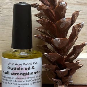 May include: A small, clear glass bottle with a black cap filled with a yellow liquid. The bottle has a label that reads "Wild Acre Wood Co. Cuticle oil & nail strengthener" and lists the ingredients: grapeseed oil, sweet almond oil, jojoba oil, vitamin E, lavender EO, geranium EO, and frankincense EO. The bottle is sitting on a wooden surface next to a brown pine cone.