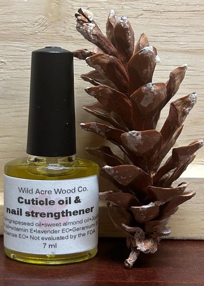 May include: A small, clear glass bottle with a black cap filled with a yellow liquid. The bottle has a label that reads "Wild Acre Wood Co. Cuticle oil & nail strengthener" and lists the ingredients: grapeseed oil, sweet almond oil, jojoba oil, vitamin E, lavender EO, geranium EO, and frankincense EO. The bottle is sitting on a wooden surface next to a brown pine cone.