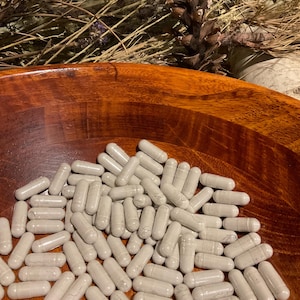 May include: A wooden bowl filled with numerous gray capsules. The capsules are oval-shaped and appear to be filled with a powder. The bowl is a rich, warm brown color, and the background includes dried plants and a pine cone.