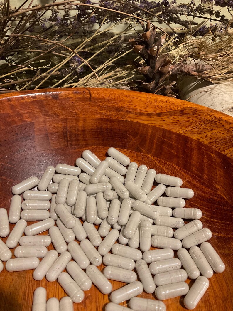 May include: A wooden bowl filled with numerous gray capsules. The capsules are oval-shaped and appear to be filled with a powder. The bowl is a rich, warm brown color, and the background includes dried plants and a pine cone.