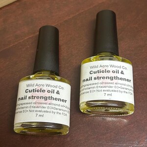 May include: Two 7 ml bottles of cuticle oil and nail strengthener by Wild Acre Wood Co. The bottles are clear glass with black caps and yellow liquid inside. The label is white with black text. The ingredients are listed on the label: grapeseed oil, sweet almond oil, jojoba oil, vitamin E, lavender essential oil, geranium essential oil, and frankincense essential oil. The label also states that the product is not evaluated by the FDA.