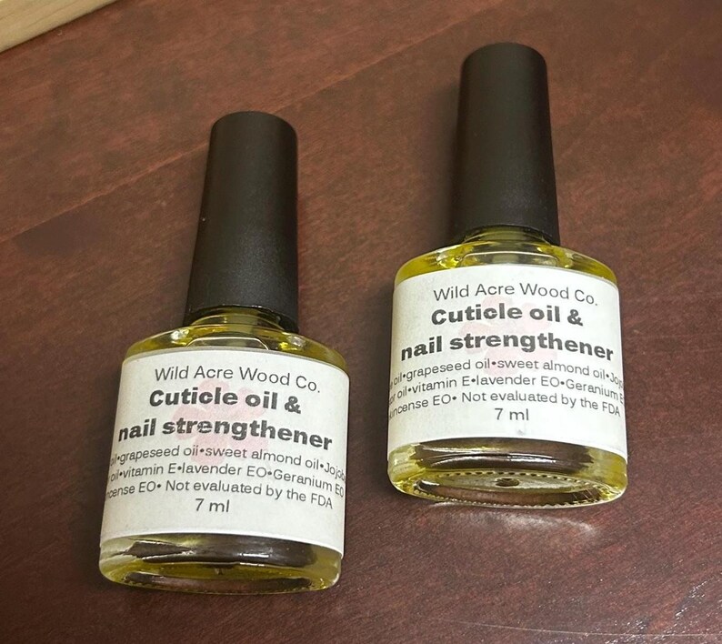 May include: Two 7 ml bottles of cuticle oil and nail strengthener by Wild Acre Wood Co. The bottles are clear glass with black caps and yellow liquid inside. The label is white with black text. The ingredients are listed on the label: grapeseed oil, sweet almond oil, jojoba oil, vitamin E, lavender essential oil, geranium essential oil, and frankincense essential oil. The label also states that the product is not evaluated by the FDA.