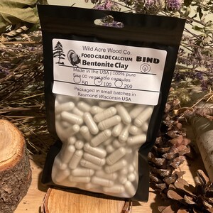 May include: A black pouch filled with size 00 vegetable capsules of Bentonite Clay. The label reads "Wild Acre Wood Co. FOOD GRADE CALCIUM BIND Bentonite Clay." The pouch is on a wooden surface with dried flowers and pine cones.