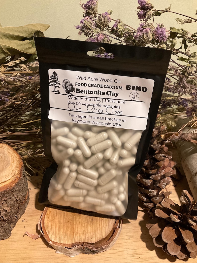 May include: A black pouch filled with size 00 vegetable capsules of Bentonite Clay. The label reads "Wild Acre Wood Co. FOOD GRADE CALCIUM BIND Bentonite Clay." The pouch is on a wooden surface with dried flowers and pine cones.