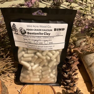 May include: A black bag filled with white capsules, labeled "Wild Acre Wood Co. Bentonite Clay." The label also states "Made in the USA" and "Size 00 vegetable capsules." The bag is surrounded by natural elements like pine cones and dried flowers.