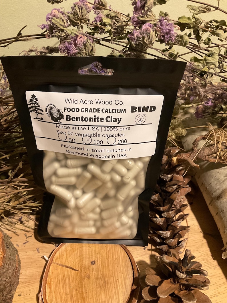 May include: A black bag filled with white capsules, labeled "Wild Acre Wood Co. Bentonite Clay." The label also states "Made in the USA" and "Size 00 vegetable capsules." The bag is surrounded by natural elements like pine cones and dried flowers.