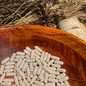 May include: A wooden bowl filled with numerous gray capsules. The bowl has a rich, reddish-brown color and a smooth, polished finish. Dried lavender and a pine cone are visible in the background, suggesting a natural or herbal theme.