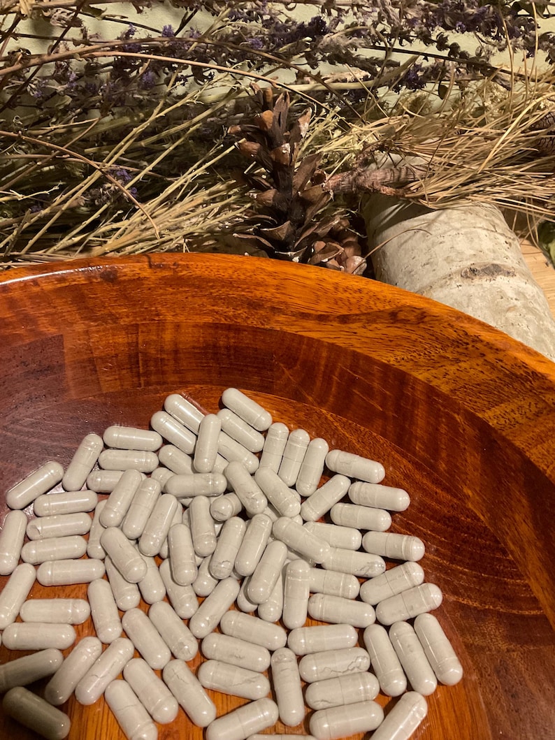 May include: A wooden bowl filled with numerous gray capsules. The bowl has a rich, reddish-brown color and a smooth, polished finish. Dried lavender and a pine cone are visible in the background, suggesting a natural or herbal theme.