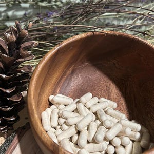 May include: A wooden bowl filled with white capsules. The bowl is sitting on a wooden surface with a pine cone to the left.