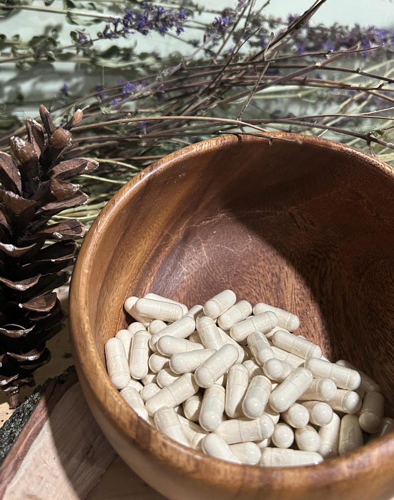 May include: A wooden bowl filled with white capsules. The bowl is sitting on a wooden surface with a pine cone to the left.