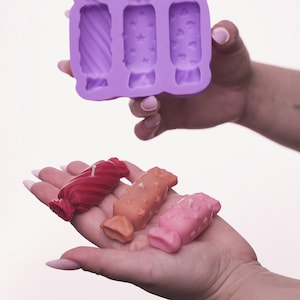 Könnte beinhalten: Eine lila Silikon-Süßigkeitenform mit drei Hohlräumen, jede mit einem anderen Design. Drei bonbonförmige Kerzen in Rot, Orange und Rosa werden in einer Hand gehalten. Die Kerzen haben ein strukturiertes Aussehen und sind angezündet.