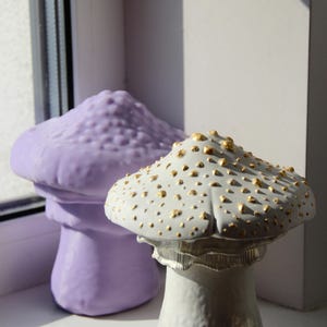 Peut inclure: Deux figurines de champignons en céramique, l'une est violette avec une surface texturée et l'autre est blanche avec des accents dorés.