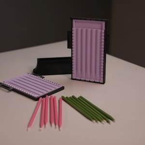 May include: A black box with a lavender insert and several pink and green candles. A second box with a lavender insert is open next to the first. A third box is open in the background. The candles are arranged in two groups.