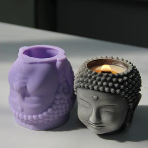 Puede incluir: Dos portavelas con forma de cabeza de Buda, uno es un molde de silicona morado y el otro es un portavelas de hormigón gris con una vela encendida dentro.