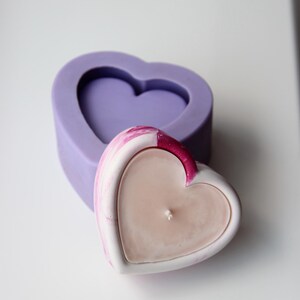 May include: A heart-shaped candle with a pink and white marbled design, sitting inside a purple silicone heart-shaped mold.