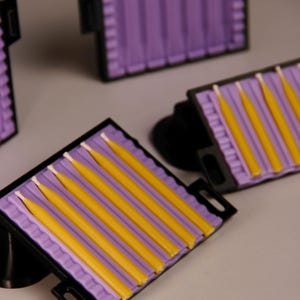 Peut inclure: Un ensemble de moules à bougies en plastique noir et violet, chacun doté de multiples canaux parallèles. Plusieurs bougies effilées jaunes avec des mèches blanches visibles reposent dans les sections moulées violettes des cadres rectangulaires.