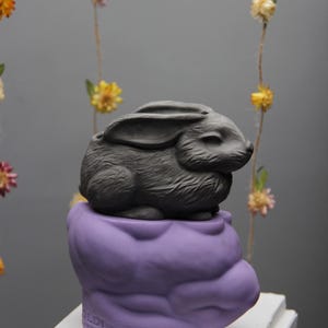 Könnte beinhalten: Ein grauer Keramik-Hasentopf steht auf einem violetten Keramik-Wolkentopf. Die Töpfe stehen auf einer weißen Holzplattform. Der Text "OLDI" und "AMOUR" ist auf der Unterseite des Wolkentopfs sichtbar.