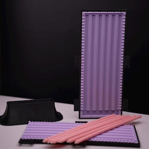 May include: A lavender silicone mold with vertical lines, a black base, and a black stand. Several pink sticks are arranged on a matching lavender tray. The background is black and white.