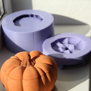Peut inclure: Un récipient en terre cuite en forme de citrouille avec un couvercle, à côté de deux moules en silicone violet pour faire des citrouilles.