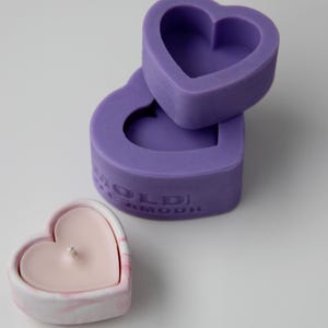 May include: Two purple heart-shaped silicone molds stacked on top of each other. The top mold has a smaller heart-shaped indentation inside. A pink heart-shaped candle is sitting on a white surface.