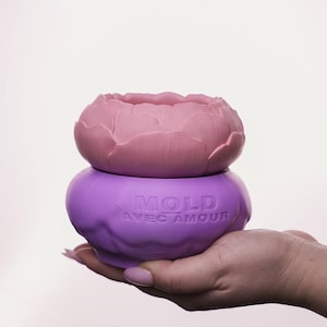 May include: A two-piece decorative container. The top piece is a light pink, flower-shaped mold. The bottom piece is a purple, rounded container with the text "MOLD AVEC AMOUR".