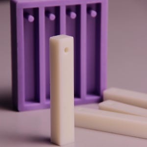 Könnte beinhalten: Nahaufnahme eines weißen, rechteckigen Stabs mit einem Loch in der Nähe der Oberseite, neben einer lila Form mit vier Schlitzen. Mehrere ähnliche Stäbe sind in der Nähe verstreut. Die Artikel scheinen zum Basteln zu sein.