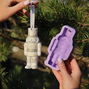 May include: A white porcelain nutcracker ornament with gold accents and a purple silicone mold of a nutcracker. The ornament is hanging from a white ribbon.