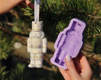 May include: A white porcelain nutcracker ornament with gold accents and a purple silicone mold of a nutcracker. The ornament is hanging from a white ribbon.