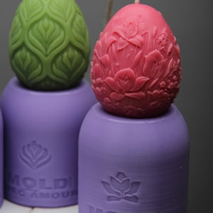 Könnte beinhalten: Zwei dekorative Kerzen in Eiform, eine grün und eine rosa, mit aufwendigen Blumendesigns. Die Kerzen stehen auf violetten Sockeln mit dem geprägten Text "Moldi Avec Amour" auf der Vorderseite.