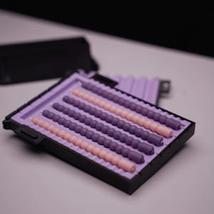 Puede incluir: Una bandeja rectangular negra contiene varias velas en espiral en tonos morados y rosas. Las velas están dispuestas en filas paralelas dentro de un inserto de color lavanda. La bandeja está sobre un fondo blanco.