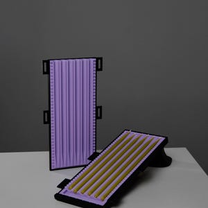 May include: Two candle molds, one upright and one angled, with a black frame and lavender interior. The angled mold contains gold-colored candles. The upright mold is empty. The background is a solid gray.