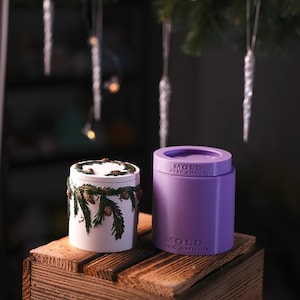 May include: A white cylindrical container decorated with green pine branches and brown pinecones alongside a purple silicone mould. The purple mould features the embossed text MOLD AVEC AMOUR on its side and top surface, indicating its function.