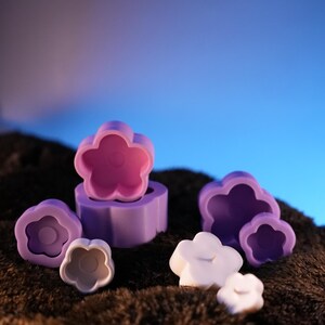 May include: A set of six purple flower-shaped silicone molds for making candles or soap. The molds are arranged on a brown fuzzy surface.