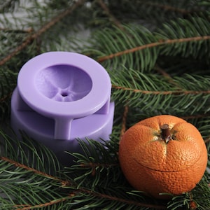 Könnte beinhalten: Ein Stillleben mit einer kleinen orangefarbenen Schmuckschatulle und einer lavendelfarbenen Form, die auf einem Bett aus grünen Tannennadeln ruht. Die orangefarbene Schachtel hat eine strukturierte Oberfläche und einen kleinen Stiel. Die Form hat eine kreisförmige Oberseite.
