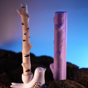 May include: A white ceramic bird with a gold leaf design holds a white birch branch. A purple branch stands next to the bird on a brown mossy surface.